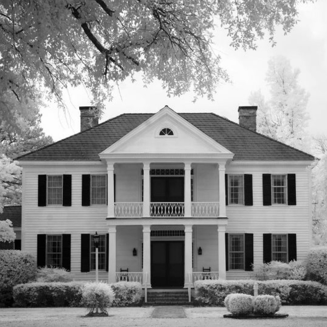 A black and white photograph of an elegant two-story colonial home with white columns and dual porches. The historic White House featured in the Bennett Auction Series by Kay Brooks stands against a misty background with overhanging tree branches. 